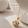 A gold rose-shaped pendant necklace with a clear central gemstone, displayed on a textured beige surface with abstract white forms in the background. Arabic text is visible near the top.