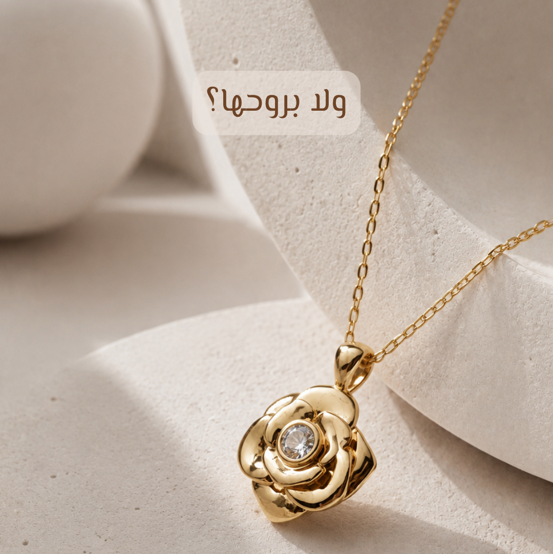 A gold rose-shaped pendant necklace with a clear central gemstone, displayed on a textured beige surface with abstract white forms in the background. Arabic text is visible near the top.
