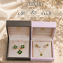 Two open jewelry gift boxes displayed on a beige fabric surface with dried flowers. The left box is dark gray and contains a gold-tone necklace and matching earrings with green stone accents.