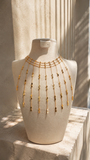 A gold-toned chain necklace with multiple draped fringe strands and small dangling disc charms, displayed on a cream-colored mannequin bust in warm natural light.