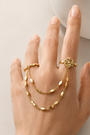 A hand wearing a coordinated gold jewelry set, including a rose-shaped ring, a delicate chain bracelet draped across the fingers, and a matching chain bracelet on the wrist, against a soft light beige background.