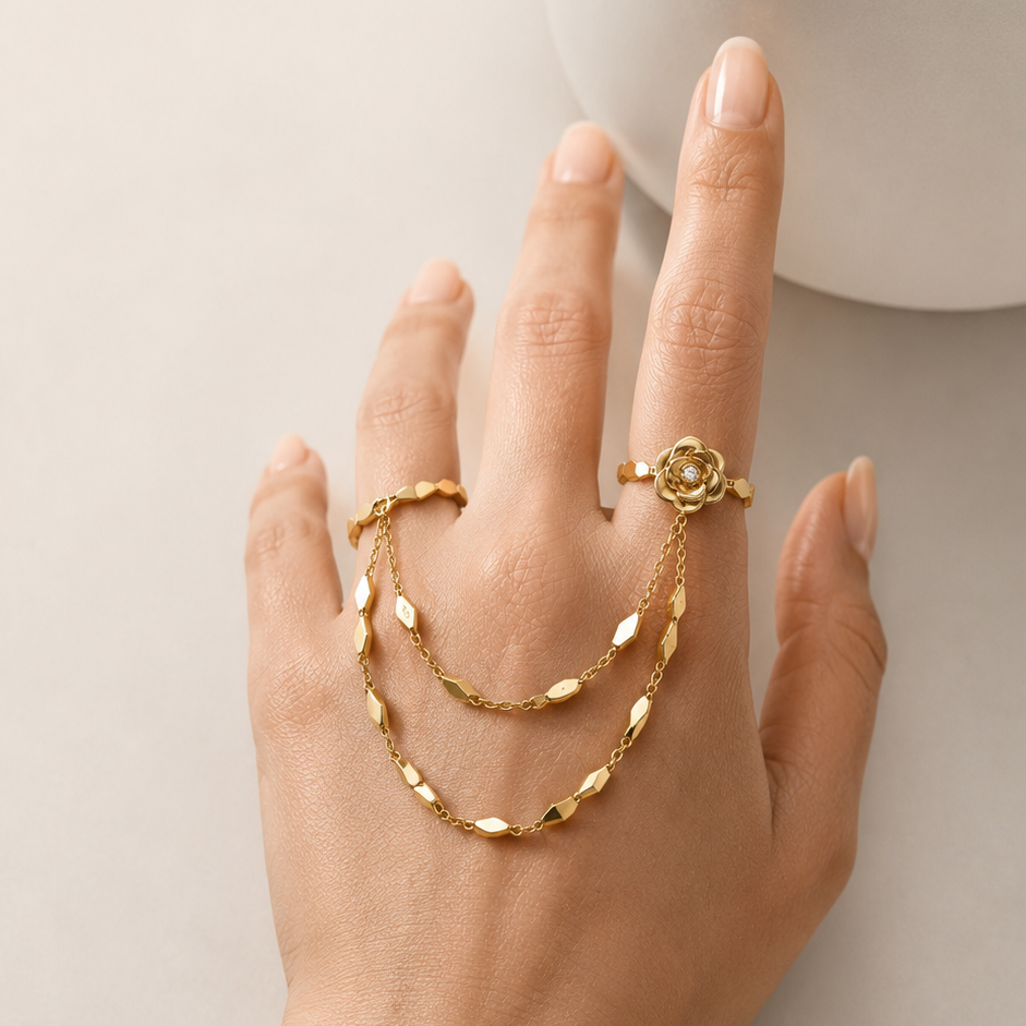 A hand wearing a coordinated gold jewelry set, including a rose-shaped ring, a delicate chain bracelet draped across the fingers, and a matching chain bracelet on the wrist, against a soft light beige background.