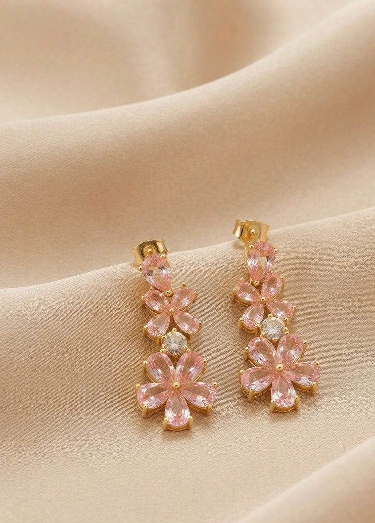 A pair of gold-tone drop earrings featuring pink flower-shaped crystals and a central clear round stone, displayed on draped beige fabric.