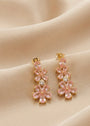 A pair of gold-tone drop earrings featuring pink flower-shaped crystals and a central clear round stone, displayed on draped beige fabric.
