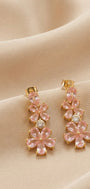 A pair of gold-tone drop earrings featuring pink flower-shaped crystals and a central clear round stone, displayed on draped beige fabric.