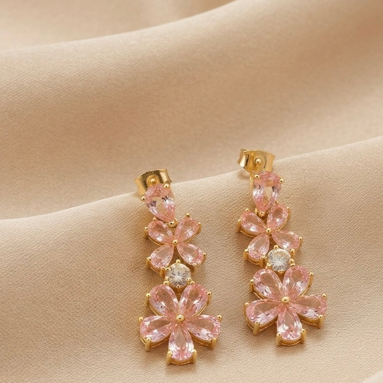 A pair of gold-tone drop earrings featuring pink flower-shaped crystals and a central clear round stone, displayed on draped beige fabric.