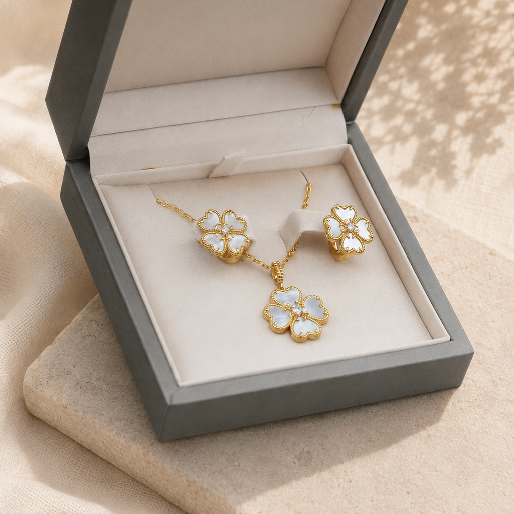 A gold-tone jewelry set with matching necklace and earrings featuring white flower-shaped stones, presented in an open gray gift box on beige fabric.