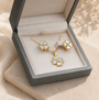 A gold-tone jewelry set with matching necklace and earrings featuring white flower-shaped stones, presented in an open gray gift box on beige fabric.
