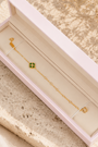 A delicate gold chain bracelet with a small green clover charm, presented in an open pale pink jewelry box on a textured beige stone slab with soft cream fabric and dried flowers in the background.