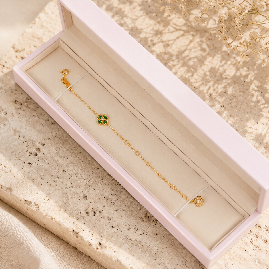 A delicate gold chain bracelet with a small green clover charm, presented in an open pale pink jewelry box on a textured beige stone slab with soft cream fabric and dried flowers in the background.