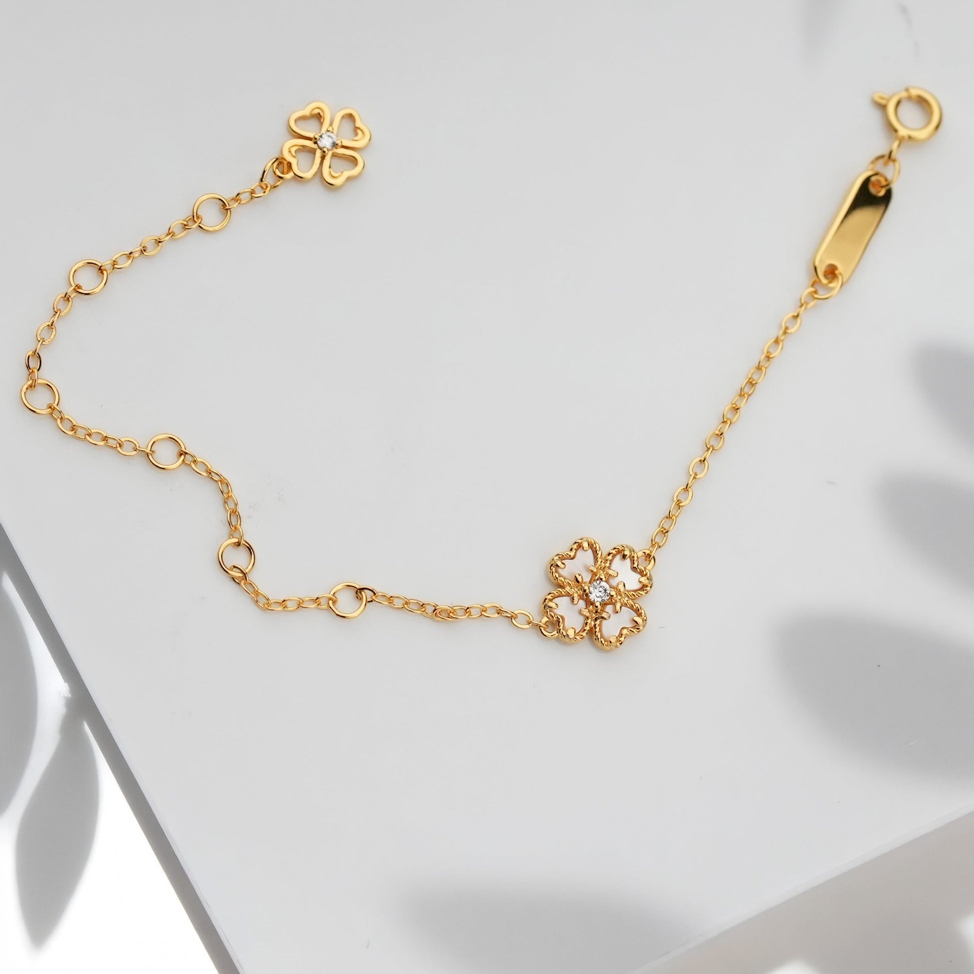 A delicate gold chain bracelet with a small four-leaf clover charm and an adjustable extender chain, styled on a white surface with soft leaf shadows.