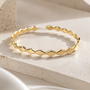 A gold geometric open cuff bracelet with a textured, faceted chevron pattern, displayed on a light beige stone block with soft white fabric in the foreground.