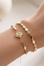 A close-up of a person's wrist wearing two delicate gold bracelets, one with a rose charm and the other with elongated gold beads, set against a soft, warm neutral background.