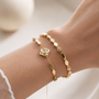A close-up of a person's wrist wearing two delicate gold bracelets, one with a rose-shaped charm and one with geometric hexagonal links, against a soft, warm neutral background.