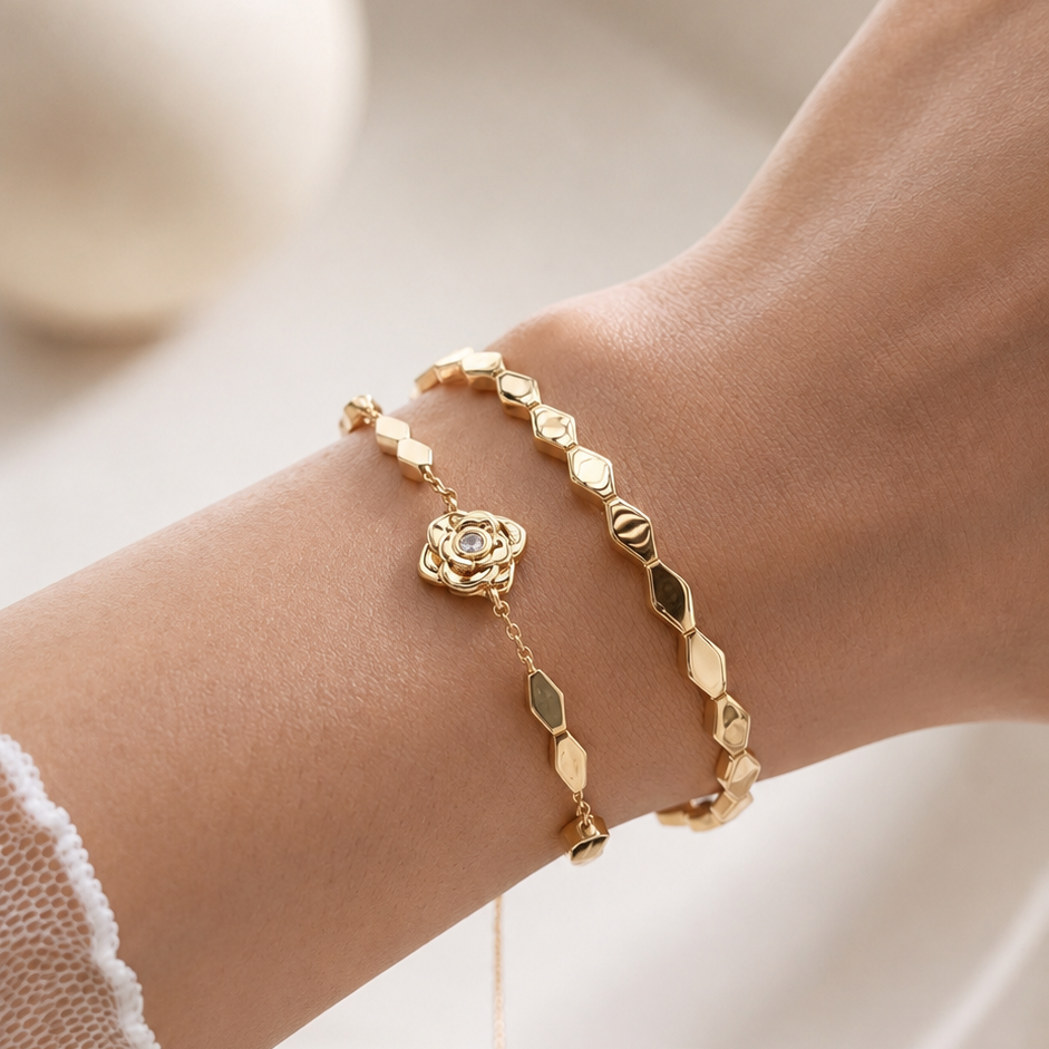 A close-up of a person's wrist wearing two delicate gold bracelets, one with a rose-shaped charm and one with geometric hexagonal links, against a soft, warm neutral background.