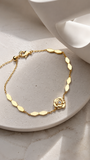 A delicate gold chain bracelet with a small rose-shaped charm and a tiny clear stone, displayed on a light beige stone surface with a white ceramic cup in the background.