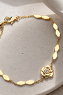 A delicate gold chain bracelet with a small rose-shaped charm and a tiny clear stone, displayed on a light beige stone surface with a white ceramic cup in the background.