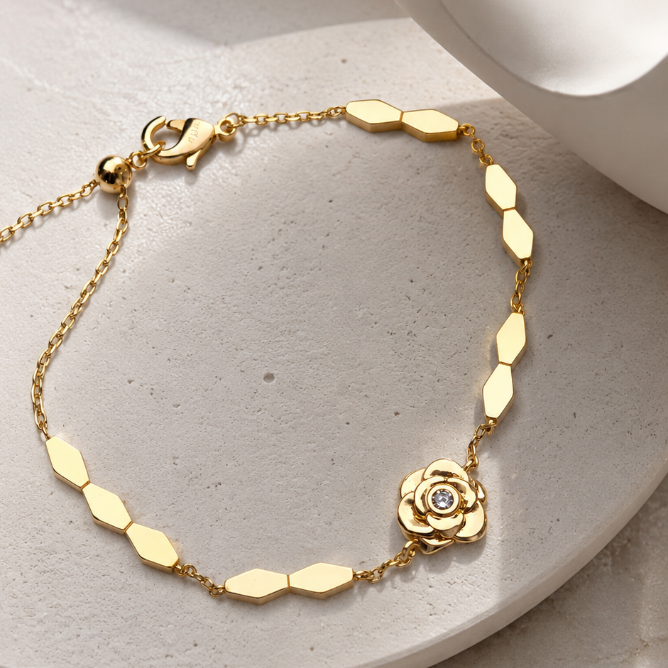 A delicate gold chain bracelet with a small rose-shaped charm and a tiny clear stone, displayed on a light beige stone surface with a white ceramic cup in the background.
