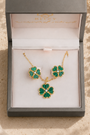 A gold-tone jewelry set with green clover-shaped stones, including a necklace and matching earrings, presented in an open gray gift box on a beige fabric surface with dried flowers.