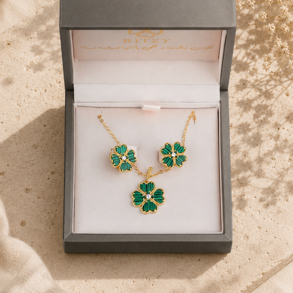 A gold-tone jewelry set with green clover-shaped stones, including a necklace and matching earrings, presented in an open gray gift box on a beige fabric surface with dried flowers.