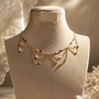 A delicate gold leaf necklace displayed on a cream-colored jewelry bust, set against a warm, softly lit background with lace and dried floral elements.