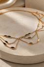 A delicate gold chain necklace with multiple layered strands and small dark oval inlays, displayed on a round beige stone pedestal with soft warm lighting.