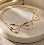 A delicate gold chain necklace with multiple layered strands and small dark oval inlays, displayed on a round beige stone pedestal with soft warm lighting.