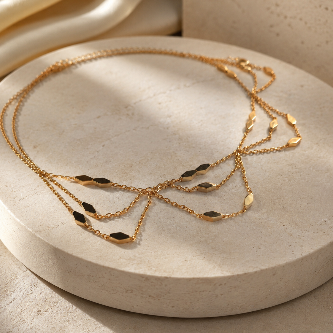 A delicate gold chain necklace with multiple layered strands and small dark oval inlays, displayed on a round beige stone pedestal with soft warm lighting.