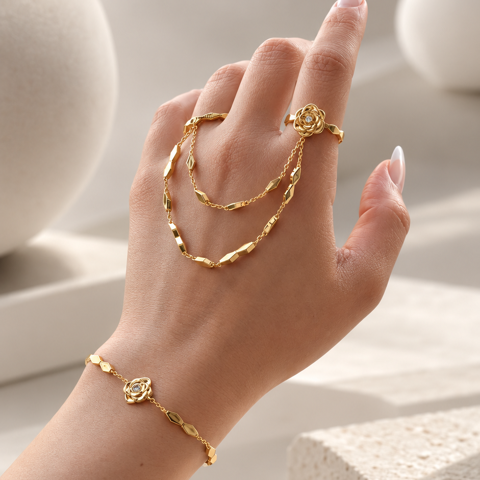 A hand wearing a delicate gold jewelry set including a bracelet, ring, and chain accessory, styled with neutral geometric props in a bright, minimalist setting.