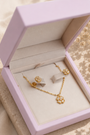 A gold jewelry set with a necklace and matching earrings displayed inside an open light lavender jewelry box, placed on a beige stone slab with soft cream fabric and dried flowers in the background.