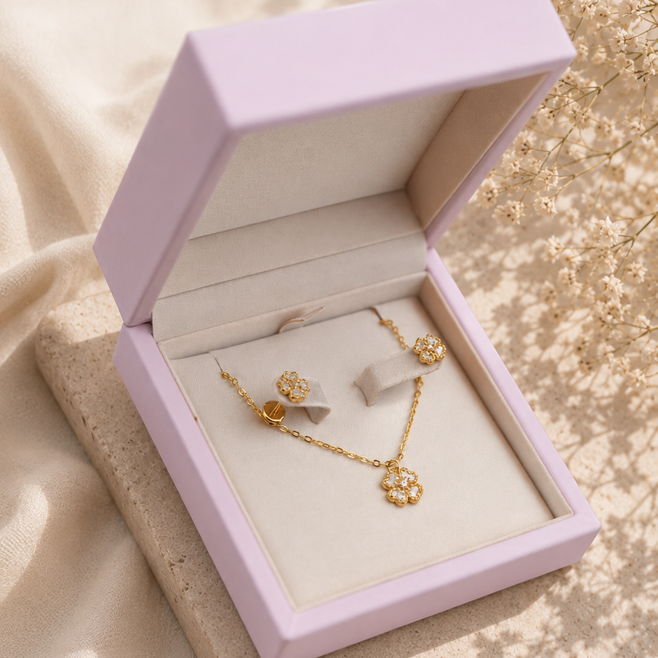 A gold jewelry set with a necklace and matching earrings displayed inside an open light lavender jewelry box, placed on a beige stone slab with soft cream fabric and dried flowers in the background.