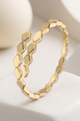 A gold geometric ring with a textured, faceted honeycomb-like design, displayed on a white sculptural stand against a soft, warm beige background.