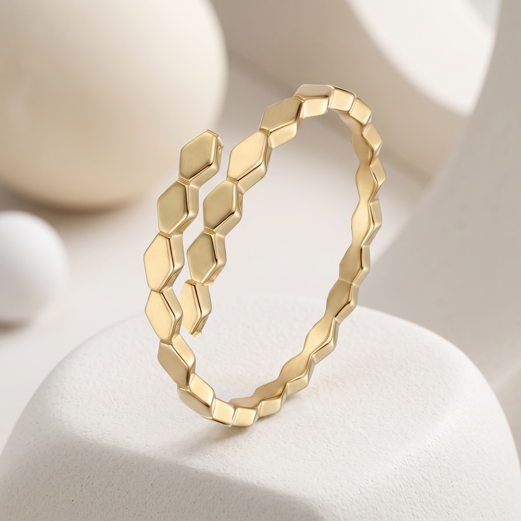 A gold geometric ring with a textured, faceted honeycomb-like design, displayed on a white sculptural stand against a soft, warm beige background.